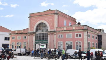 Pink building with arched windows, labeled 'Museu do Fado', people and vehicles in front on a cloudy day.