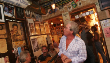 Man sings in a cozy, memorabilia-filled room with others playing guitar.