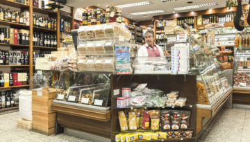Man behind counter in a store with various packaged goods and wine bottles on shelves.