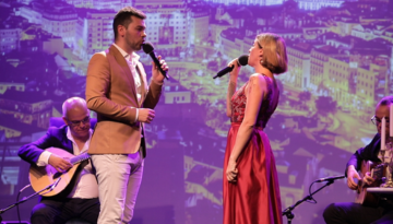 Two singers performing on stage with cityscape background; seated musicians playing guitars.