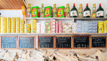 Shelves with canned fish, olive oil bottles, prices on chalkboards below.