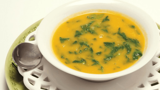 Yellow soup with green herbs in a white bowl, on a green plate with a spoon.
