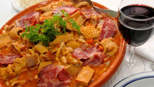 A dish of tripe stew with beans and parsley garnish beside a glass of red wine.