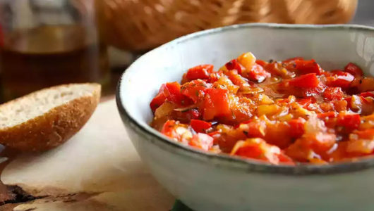 Bowl of tomato and pepper soup with bread slice on a wooden surface.