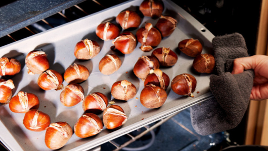 Baking tray of roasted chestnuts being held in an oven by a gloved hand.