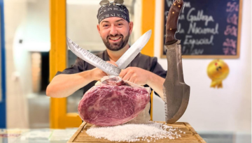 Smiling butcher holding meat with two knives crossed in the background.