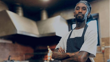 Chef in striped apron stands confidently in a restaurant kitchen with arms crossed.