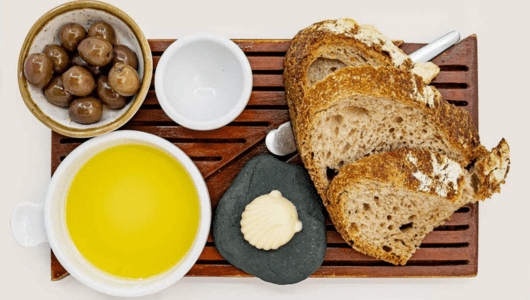 Wooden tray with olives, olive oil, butter, and bread slices.