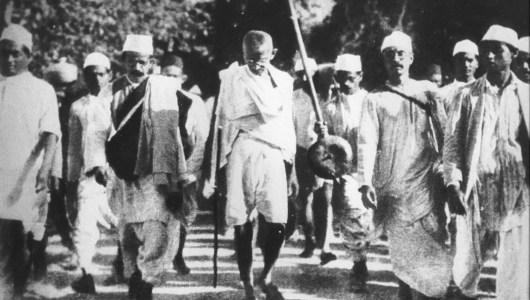 A historical black and white photo of people marching outdoors, led by a man with a walking stick.