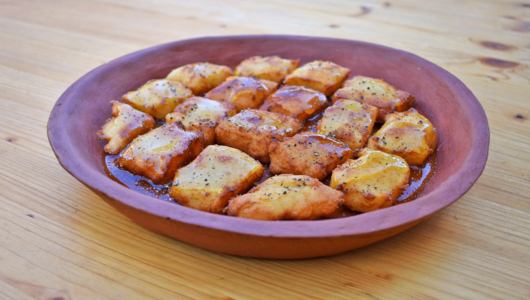 Clay dish with baked bread pieces topped with cheese and herbs on a wooden table.
