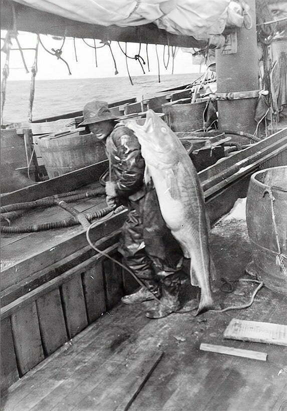 Fisherman on a boat holding a large fish with nets and barrels in the background.