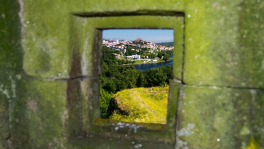 View of a town and river through a small stone window, framed by greenery.