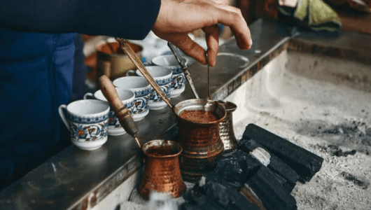 Hand making coffee in a cezve on sand with several traditional cups nearby.