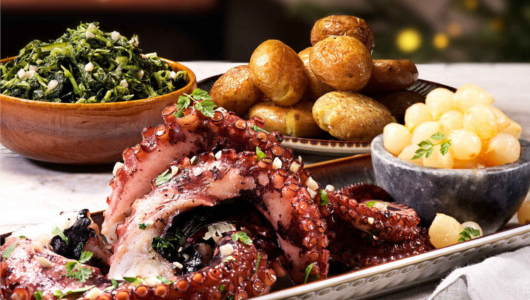 Grilled octopus with potatoes, greens, and pickled onions on a table.