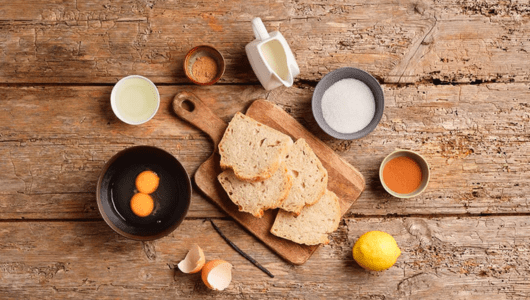 Ingredients on wooden table: bread slices, eggs, lemon, milk, sugar, spices.