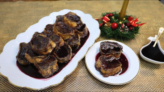 Two plates with brown pastries covered in dark sauce beside a small wreath and sauce bowl on a table.