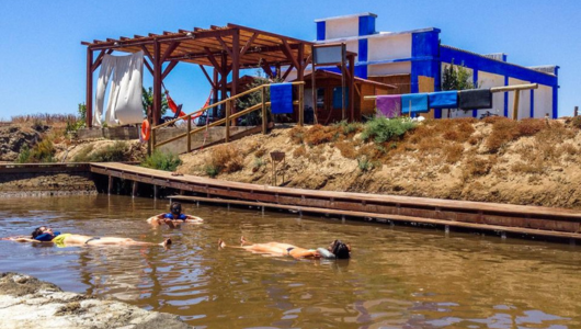 People floating in a mud pool near a blue and white building with a wooden canopy.