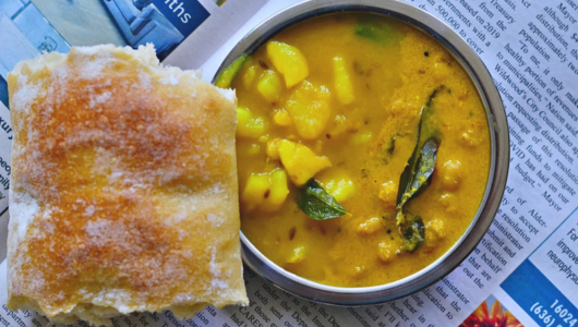 Bun and bowl of yellow curry with potatoes on a newspaper.