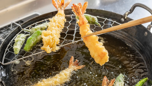 Shrimp and vegetable tempura frying in a black pan with tongs and a wire rack.