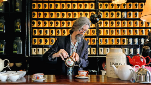 Man with grey hair pours tea in a shop with shelves of tea containers.