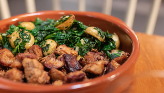 Bowl of sautéed kale, roasted potatoes, and seasoned pork chunks on a wooden table.