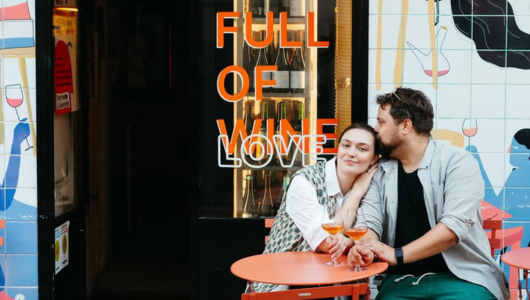 Couple sits outside a bar with an orange table, under a sign reading 'Full of Wine Love.'