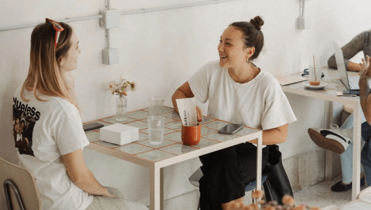 Two women smiling and talking at a cafe table with drinks and a laptop nearby.