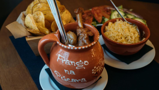 Ceramic pot with chicken, bowl of rice, basket of chips, and salad on wooden table.
