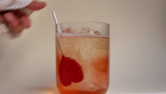 A glass of bubbly drink with a red heart stirring stick, hand reaching in.