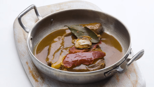 Metal pan with beef stew, bay leaves, and red pepper on cutting board.