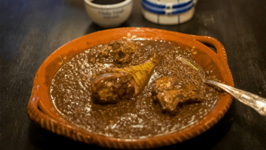 Brown stew with meat in a clay dish, a spoon, cups in the background on a dark table.