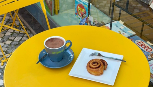 Yellow table with hot chocolate in blue cup and cinnamon roll on square plate.
