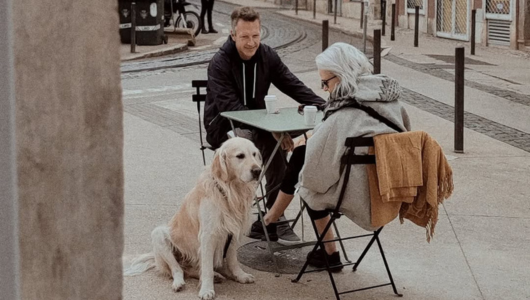 Two people sitting at an outdoor table with a dog lying beside them.
