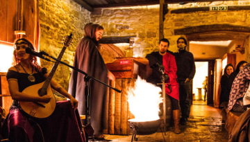 Medieval-themed gathering with musicians and people in period clothing around a fire in a stone-walled room.