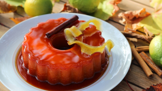 A flan dessert garnished with cinnamon sticks and lemon zest on a white plate surrounded by limes and leaves.