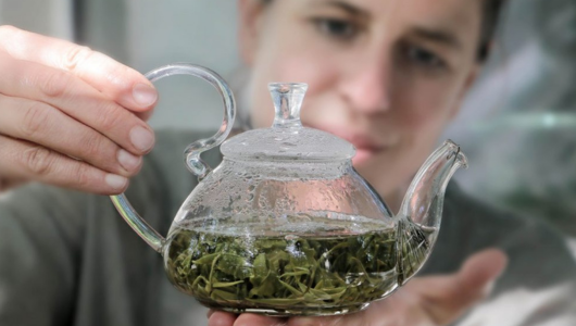 Person holding a glass teapot with green tea leaves inside.