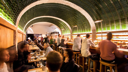 People dining and drinking at a bar with bottle-lined arched ceiling.