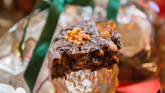 Close-up of a slice of chocolate nut cake on a glass stand with a green ribboned gift package in the background.