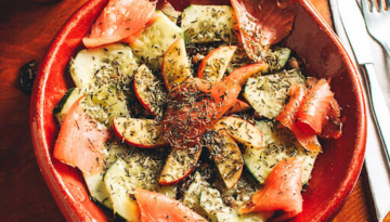 Salad with sliced smoked salmon, zucchini, apples, and herbs in a red bowl on a wooden table.