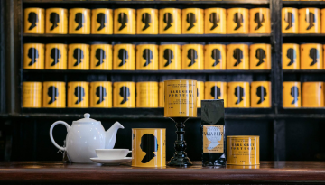 Tea products on a table with yellow tins featuring black silhouettes on shelves in the background.