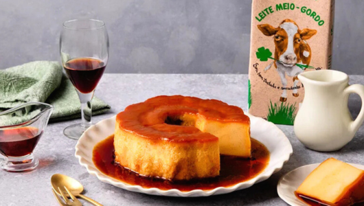 Flan dessert on plate with caramel, wine glass, milk carton, and a green napkin on a gray table.
