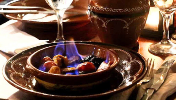 Clay dish with flaming chorizo on a dining table with wine glasses.