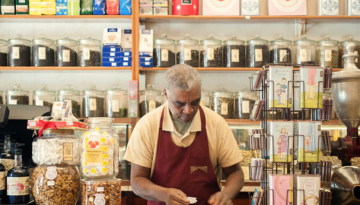 Man wearing an apron in a shop with jars and packaged goods on shelves.