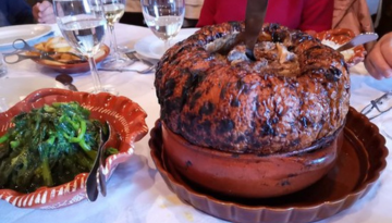 Large baked dish with knife, side of greens in brown dish on a table with wine glasses.