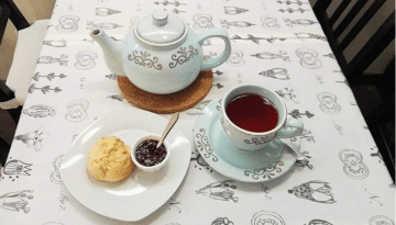 Tea set with blue teapot, cup, biscuit, and jam on a patterned tablecloth.