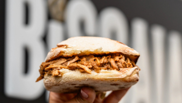 Hand holding a pulled pork sandwich against a blurred dark background.