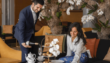 Man pours champagne for woman at a table with a tiered tray of pastries and flowers.