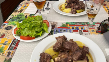 Table with plates of meat and potatoes, side salad, and beer glasses on a colorful tablecloth.