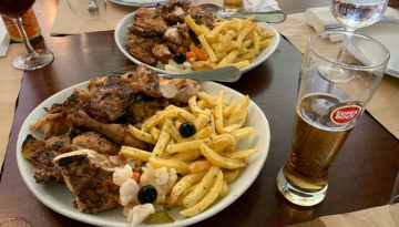 Plates of grilled chicken, fries, and pickles with a glass of beer on a table.