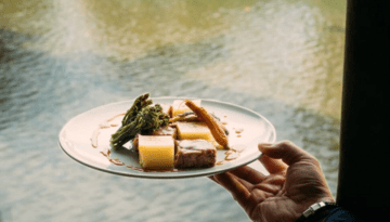 Hand holding a plate with steak, asparagus, and potatoes by a water view.